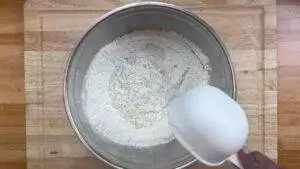 Bowl of flour with water being poured from a white measuring cup on a wooden surface. Perfect for baking or cooking.