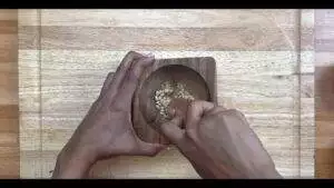 Person crushing pine nuts in a wooden mortar with a pestle on a wooden surface.