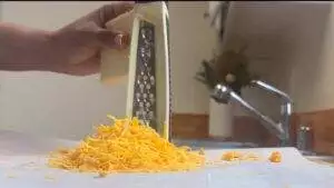 Hand grating cheddar cheese in a kitchen, with a sink and plant visible in the background.