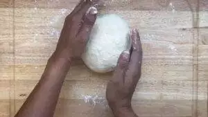 Hands kneading round dough on a floured wooden board, preparing for baking.
