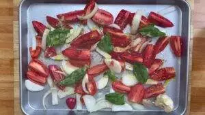 Sliced tomatoes, onions, garlic, and basil with olive oil on a baking sheet, ready for roasting.
