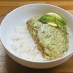 Salmon with pesto sauce on rice, garnished with basil and lemon, served in a white bowl on a wooden table.