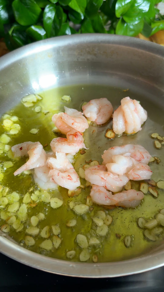 Shrimp and sliced garlic sizzling in olive oil in a stainless steel pan, with green leaves in the background.