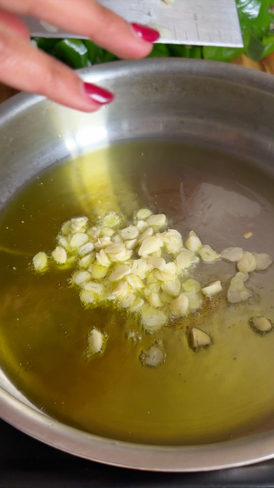 Sliced garlic being added to hot olive oil in a pan, with a hand holding a knife above the pan.