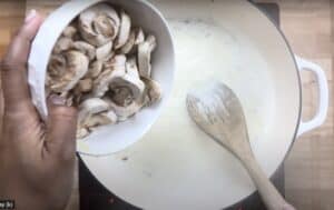 Adding sliced mushrooms to a creamy sauce in a white pan for cooking.