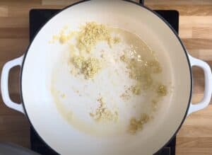 Chopped garlic sautéing in olive oil in a white pan on a wooden countertop.