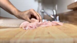 Person slicing raw chicken on a wooden cutting board in a kitchen.