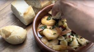 Hand dipping bread into a bowl of shrimp garlic oil tapas with herbs, served with crusty bread on a wooden table.