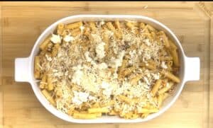 Baked ziti with cheese topping in a white dish on a wooden board, ready for serving.