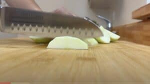 Close-up of a knife slicing green apple wedges on a wooden cutting board in a kitchen setting.