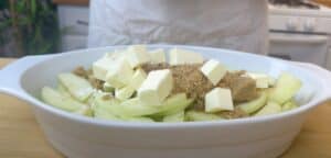 Sliced apples in a dish topped with butter and brown sugar, ready for baking. Ideal for an apple pie or dessert.
