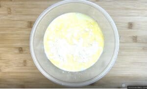 Mixing flour and eggs in a clear bowl on a wooden table, viewed from above.