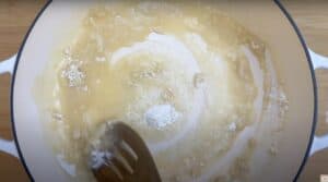 Stirring flour and butter to make a smooth roux for sauce in a white pan on a wooden surface.