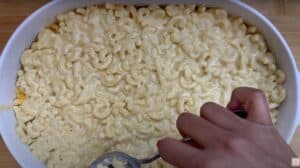 Hand scooping cheesy baked macaroni from a white casserole dish on a wooden surface.