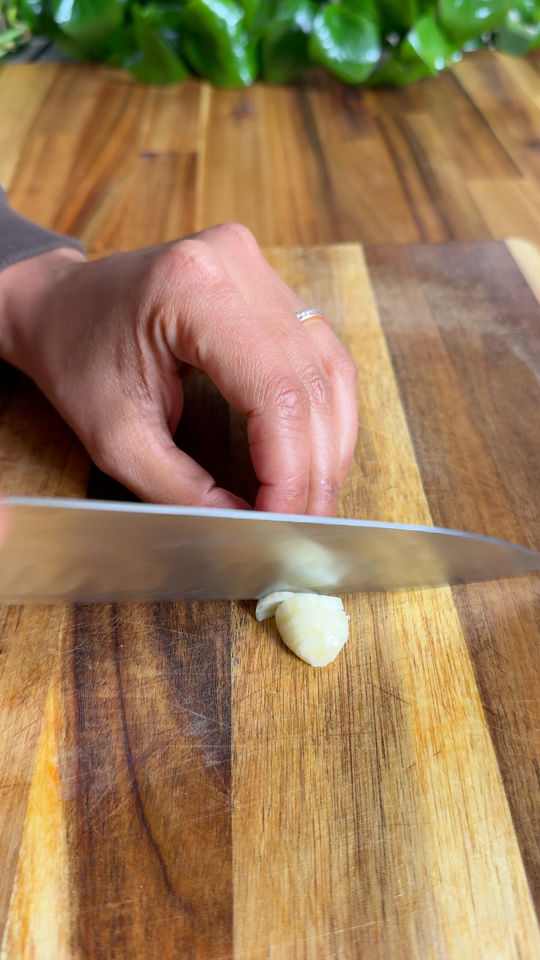 Hand slicing garlic clove on wooden cutting board.