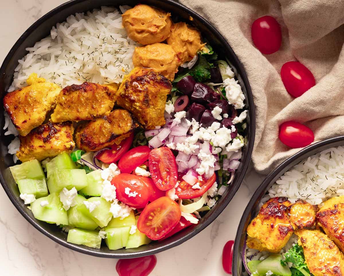 Chicken Mediterranean bowl with rice, cucumbers, cherry tomatoes, onions, feta, olives, and hummus on a white surface.
