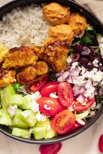 Chicken Mediterranean Bowl with grilled chicken, rice, hummus, cucumber, tomato, onion, feta cheese, beets on a marble surface.