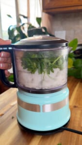 Turquoise food processor blending fresh parsley and other ingredients on wooden counter.