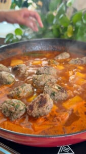 Meatballs simmering in tomato sauce with vegetables in a pan, hand adjusting heat.
