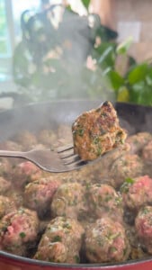 Fork holding a cooked meatball over a pan of steaming, herb-seasoned meatballs, with leafy plants in background.