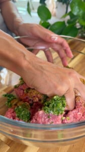 Hands mixing raw meat with herbs in a glass bowl for meatballs on a wooden countertop.