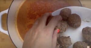Hand placing meatballs into a pot of simmering tomato sauce for cooking.