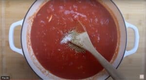 Pot of tomato sauce with herbs and salt being stirred with a wooden spoon on a wooden board.