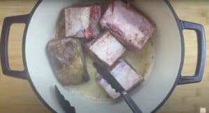 Browning beef short ribs in a Dutch oven with tongs on a wooden cutting board background.