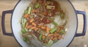 Vegetable stew prep: onions, carrots, celery, and tomato in a pot, ready for cooking on a wooden board.