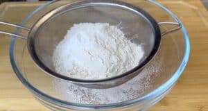 Sifting flour through a fine mesh sieve into a glass bowl on a wooden surface for baking preparation.