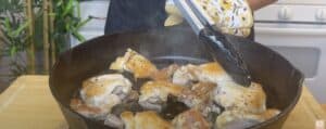 Person cooking seasoned chicken thighs in a cast iron skillet using tongs.