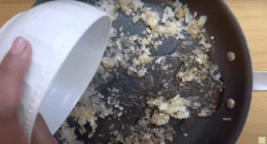 Pouring water into a sizzling pan of sautéed chopped onions on a wooden surface.