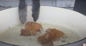 Crispy chicken breasts cooking in creamy garlic sauce, held by tongs in a white pan.