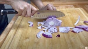Person slicing red onion on a wooden cutting board with a knife.