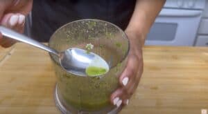 Person holding a spoon of homemade green herb sauce above a blender container on a wooden cutting board.