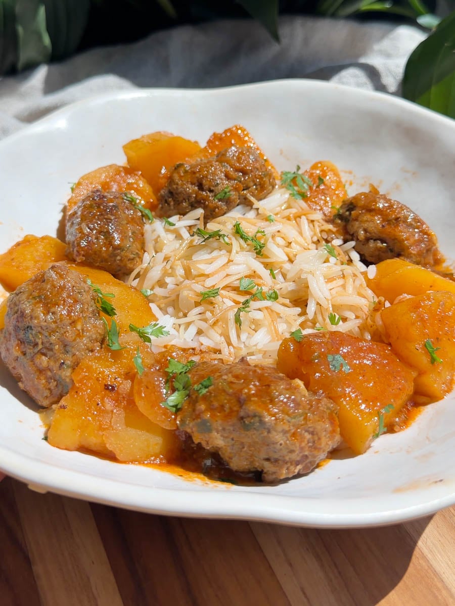 Plate of Middle Eastern meatball stew with rice in tomato sauce and roasted potatoes garnished with fresh herbs.