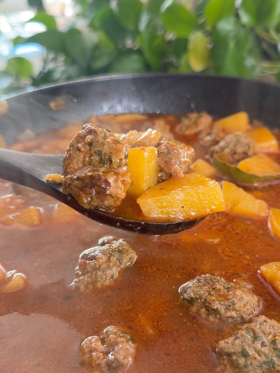 Steaming meatball and potato soup in a black ladle with fresh greens in background.