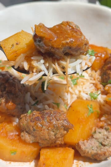 Close-up of a spoon with rice, potato, and meatball in tomato sauce garnished with parsley.