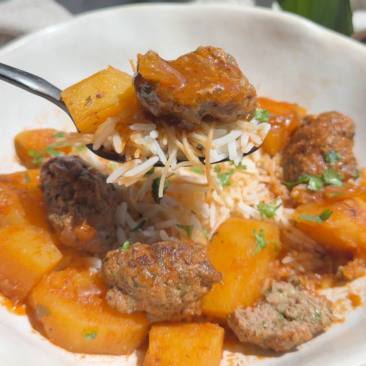 Close-up of a spoon with kofta and rice, potato, and meatball in tomato sauce garnished with parsley.