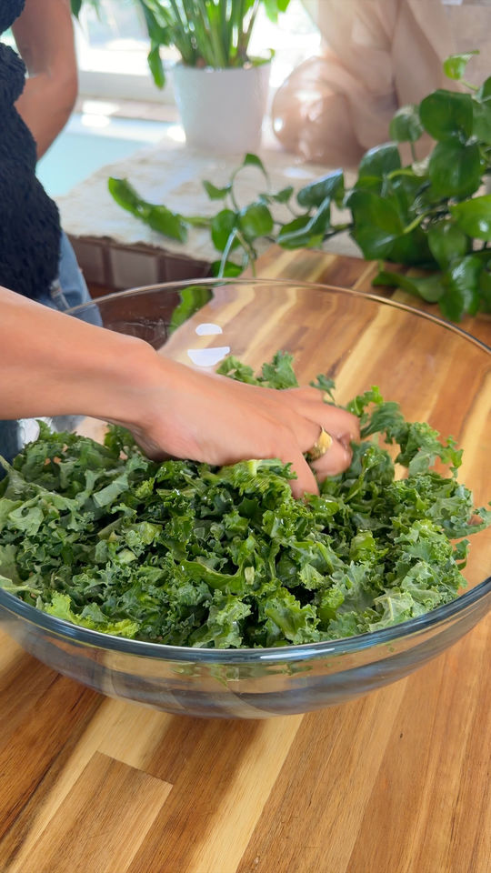 Hand mixing fresh kale in a clear glass bowl on a wooden countertop.
