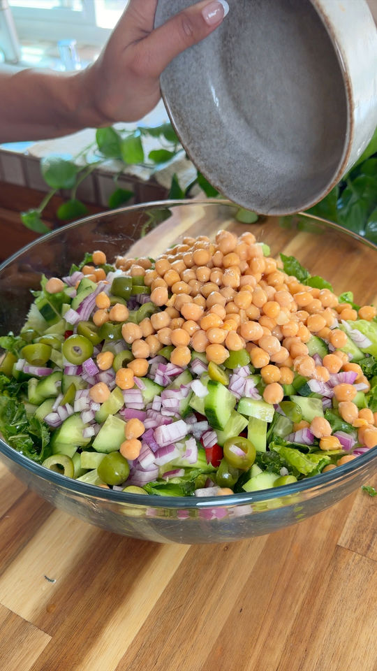 Large glass bowl with mixed salad ingredients, topped with chickpeas, being poured from a gray bowl.