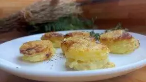 Golden cheesy potato stacks on a white plate with herbs in the background.