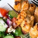 Grilled chicken skewers with a fresh cucumber tomato salad on a white plate.