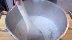 Stirring creamy broth with herbs in a stainless steel pot using a wooden spoon, cooking preparation in the kitchen.