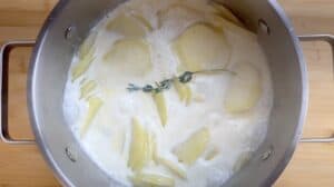 Pot of sliced potatoes simmering in creamy milk with a sprig of thyme.