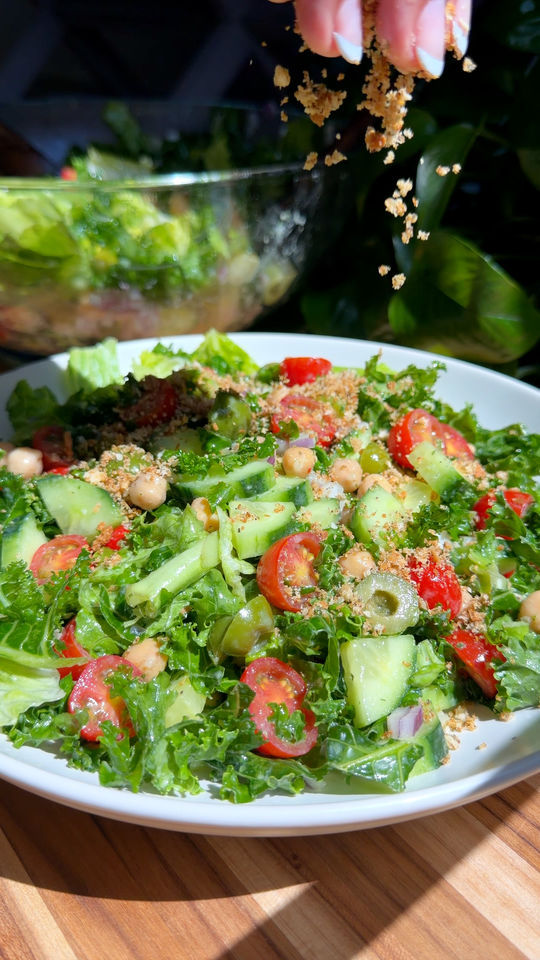 Hummus crunch salad with cherry tomatoes, cucumbers, and chickpeas being sprinkled with breadcrumbs from above.