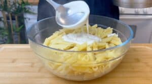 Creamy sauce being poured over a bowl of penne pasta on a wooden surface.