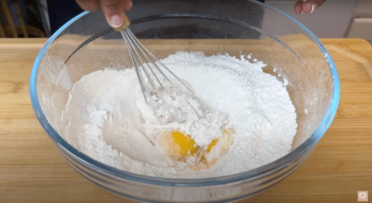 Mixing eggs and flour in a glass bowl with a whisk on a wooden surface. 