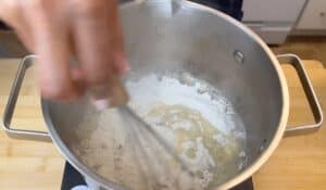 Person whisking flour and butter in a pot on a wooden counter, preparing a roux.