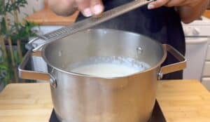 Person grating nutmeg over a simmering pot of creamy sauce in the kitchen.